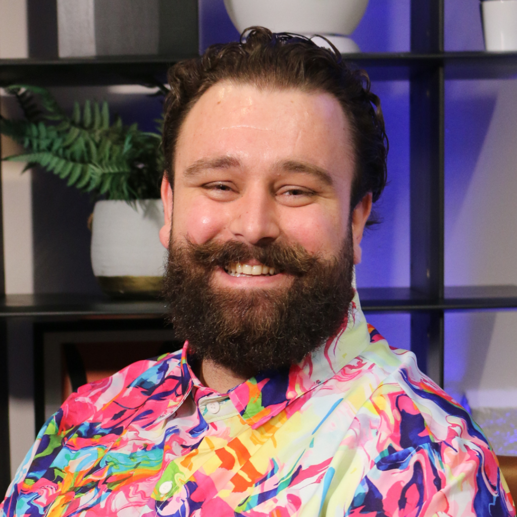 Smiling man with a full beard and moustache wearing a colourful abstract shirt, seated in front of modern shelving with plants and ceramics. The vibrant outfit and warm expression create a confident, approachable look suitable for a professional or creative headshot.